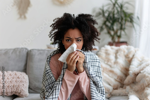 Young sick woman with cold or flue sitting on grey couch, holding a tissue to clear, blowing runny nose. Sneezing Girl under gray plaid blanket with handkerchief to dripping nose, blowing snot at home