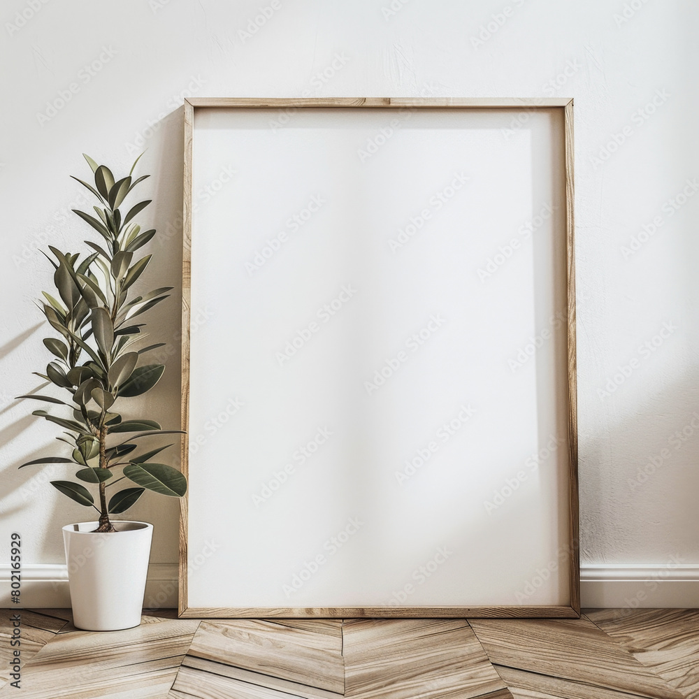 A white framed picture sits on a wooden floor next to a potted plant. The empty frame and the plant create a sense of calm and serenity in the room