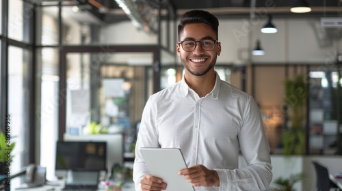 Smiling professional young latin business man company employee, male corporate manager, businessman office worker looking at camera holding digital tablet standing in office, vertical portrait