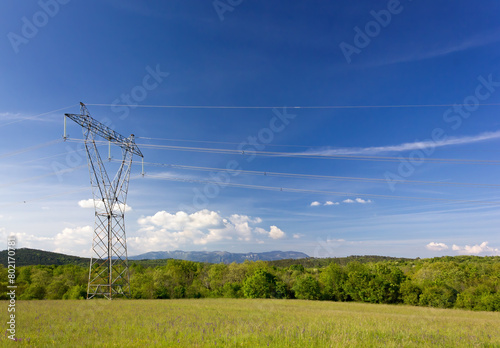 Green field against a blue sky with an electricity pylon in the foreground and hills and mountains in the background