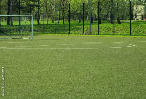 an artificial turf soccer field surrounded by a metal fence with a circle in the middle and a soccer goal on the left