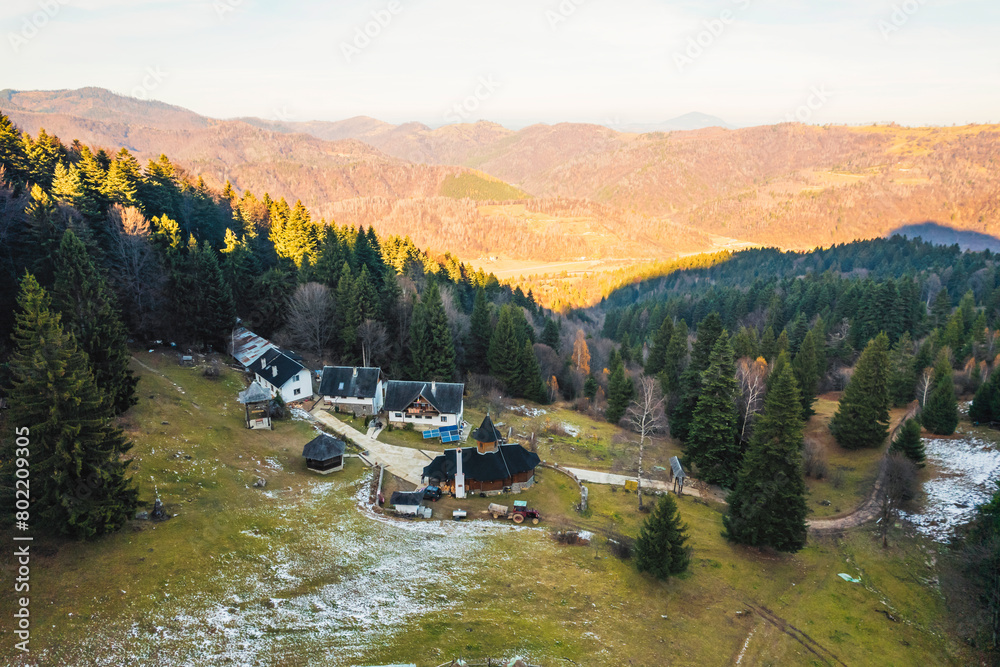 Fototapeta premium A stunning aerial view of a house nestled among the majestic mountains, surrounded by lush greenery and towering peaks under a clear blue sky