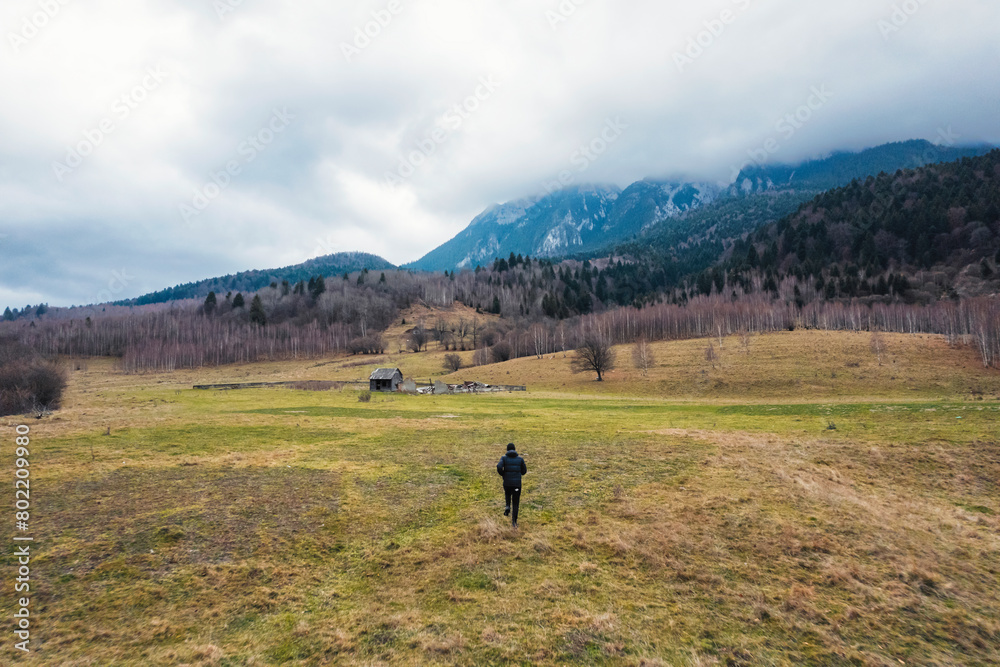 A lone figure stands in a wide field, surrounded by towering mountains in the distance. The vast expanse of nature creates a sense of peace and tranquility