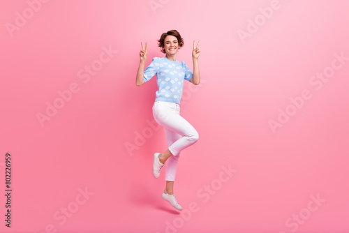 Full length photo of energetic excited person jumping demonstrate v-sign empty space isolated on pink color background