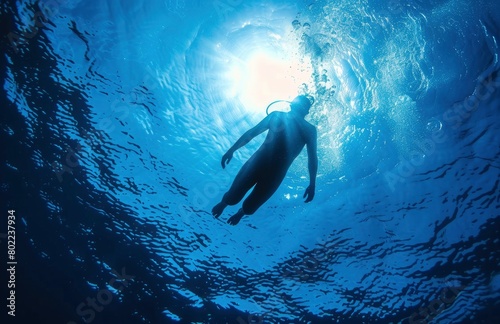 A person swimming underwater, with the sun shining coming on top of water surface.