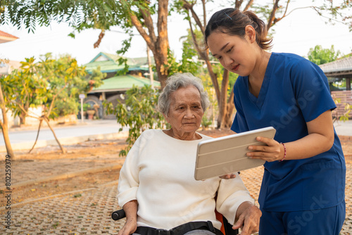 Young nurse and elderly asian senior woman on wheelchair, Asian careful caregiver teach patient do stretches to exercise arm mucsel with digital tablet at the garden on the evening. Health insurance.