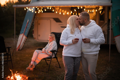 A married couple with glasses of wine stands against the background of a motorhome and rests together by the campfire. Evening family vacation photo
