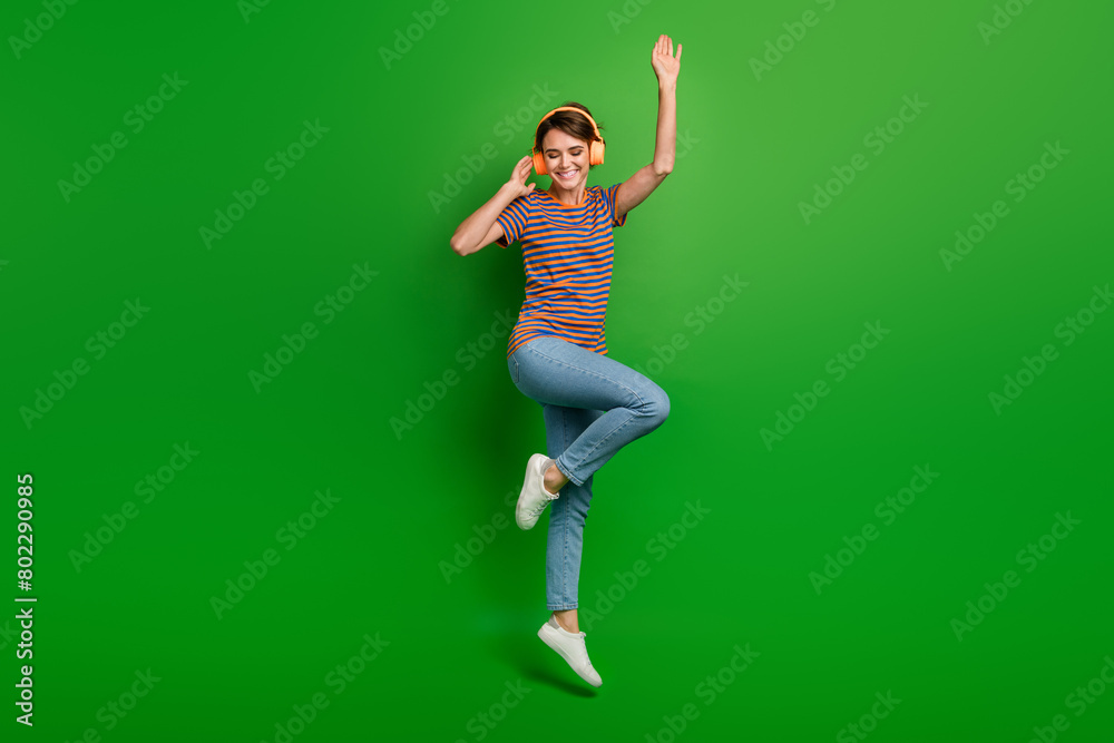 Full size portrait of cheerful lovely girl jumping hand touch headphones enjoy song isolated on green color background Full size portrait of cheerful lovely girl jumping hand touch headphones enjoy song isolated on green color background