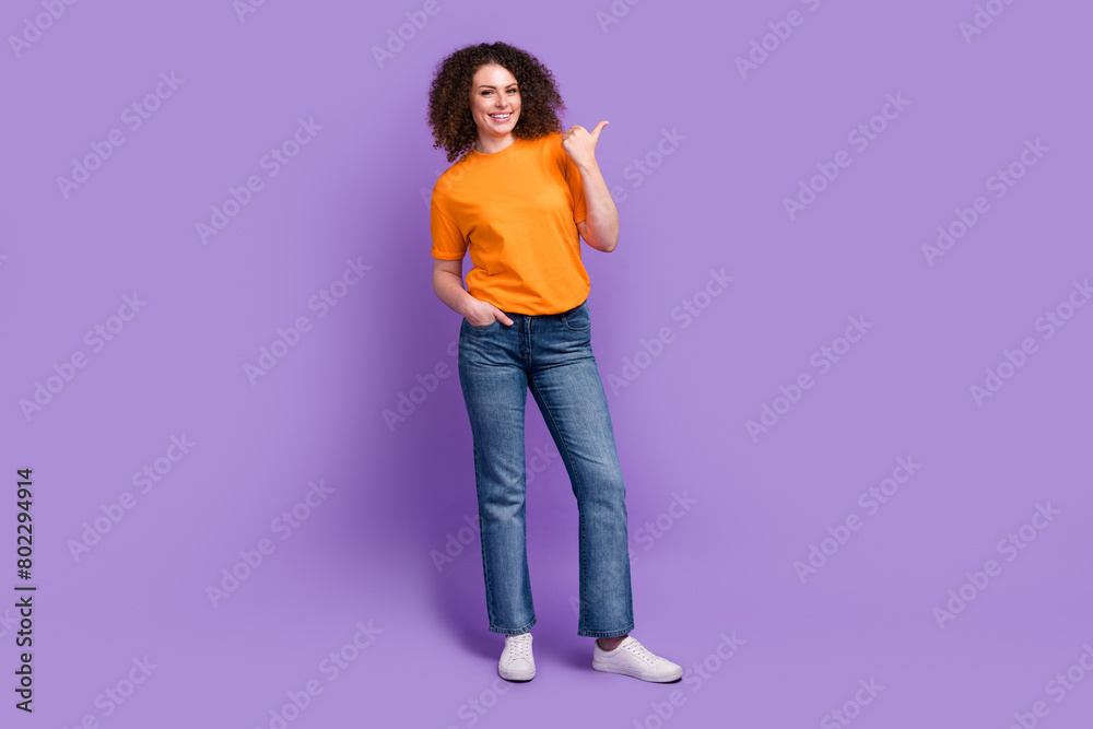 Full length photo of pretty sweet lady dressed orange t-shirt pointing thumb empty space isolated violet color background Full length photo of pretty sweet lady dressed orange t-shirt pointing thumb empty space isolated violet color background
