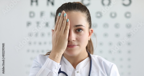 Portrait of gorgeous woman ophthalmologist standing in clinic office and covering right eye with tender hand. Lady looking at camera with happiness. Eyesight check concept photo