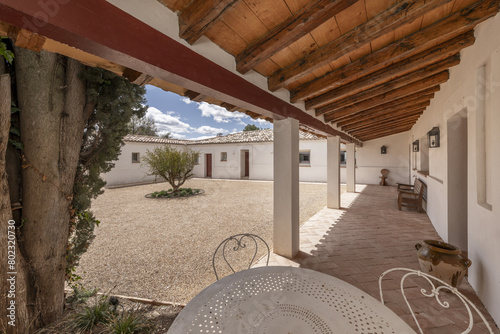 Covered porch with wooden roof of an Andalusian farmhouse style country house with terracotta sidewalks and gravel driveway.
