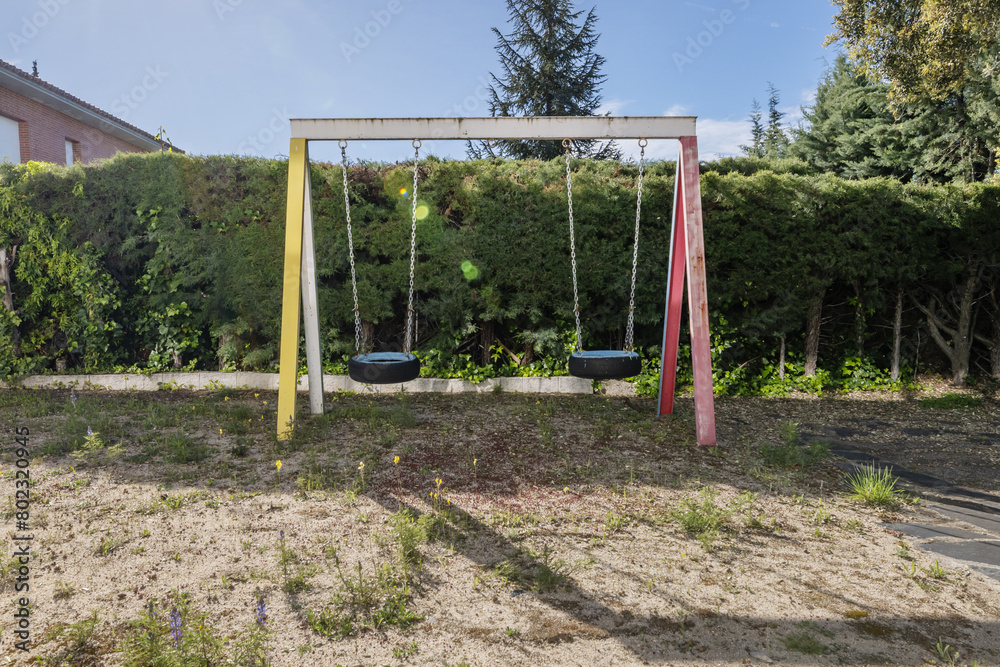Perimeter wall with hedges and children's swings with wheels as seats and sand floors with wild vegetation