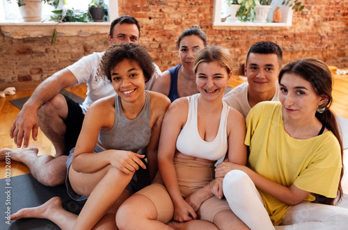 Multiracial smiling people sit on mats after doing yoga at fitness club. Group of young athletes of different age and nationalities relax and rest after physical exercises in the gym.