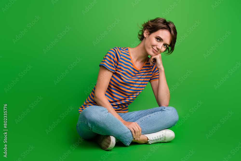 Full size portrait of lovely cheerful girl sit floor crossed legs hand touch chin isolated on green color background Full size portrait of lovely cheerful girl sit floor crossed legs hand touch chin isolated on green color background