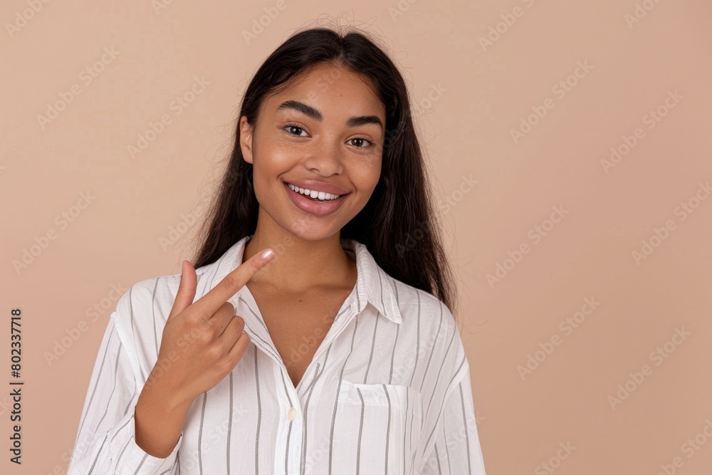 A woman smiling and pointing to the side with her right hand, wearing a white vertically striped shirt, standing in front of a solid background A woman smiling and pointing to the side with her right hand, wearing a white vertically striped shirt, standing in front of a solid background