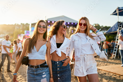 Three young women walk together on sunny beach at music festival. Happy friends in trendy summer outfits enjoy live concert, dancing, laughing in festive atmosphere. Group of girls having fun by sea.