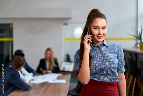 Asian businesswoman in smart casual attire talks on phone, confident, with office colleagues engaged in work behind her, embodying global connectivity, professional communication in corporate setting.