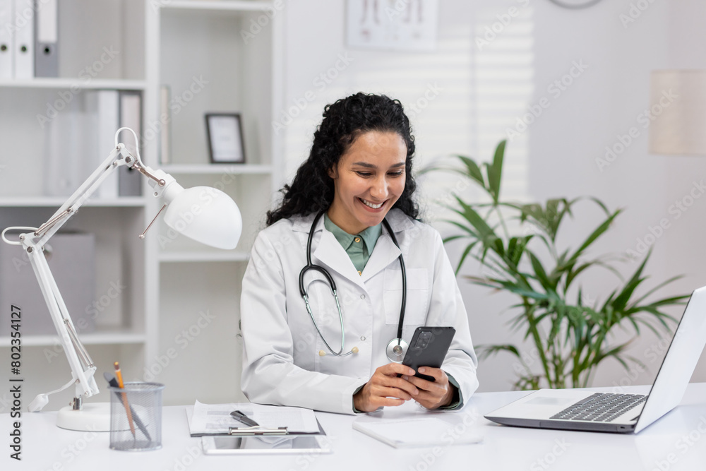 Dark haired nurse in professional uniform looking at mobile telephone screen while sitting by desk with professional equipment. Arab woman texting online while waiting for invited patient. Dark haired nurse in professional uniform looking at mobile telephone screen while sitting by desk with professional equipment. Arab woman texting online while waiting for invited patient.