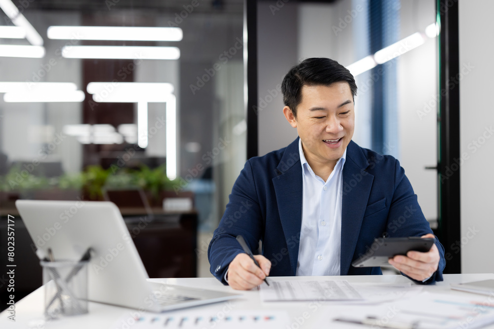 Smiling chinese accountant summarising expenses and income while using calculator and writing down indicators. Attentive economist filling document with tables during busy day in office. Smiling chinese accountant summarising expenses and income while using calculator and writing down indicators. Attentive economist filling document with tables during busy day in office.