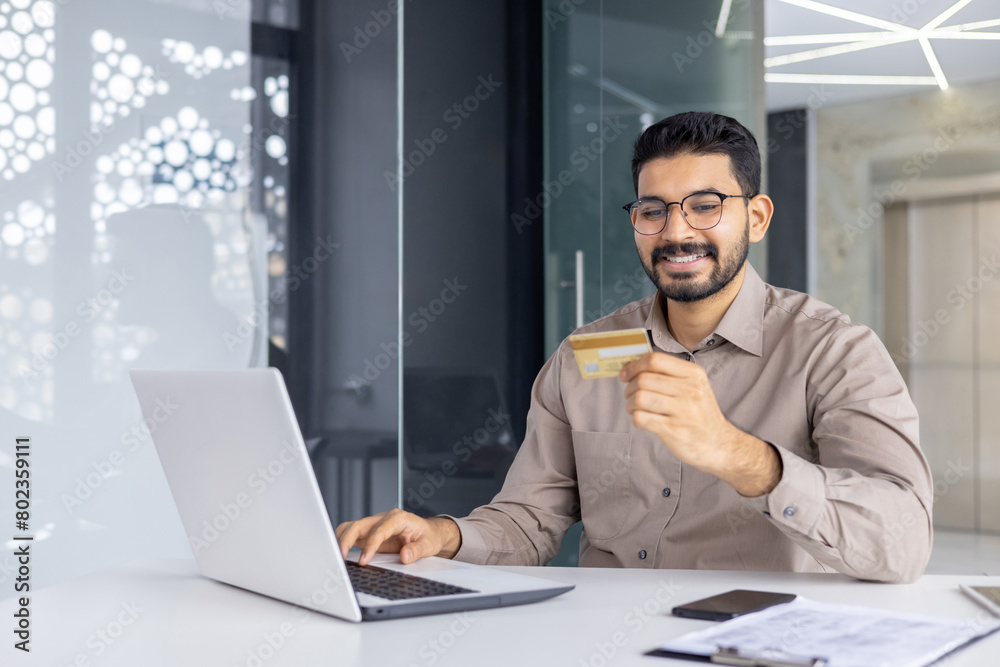 Fototapeta premium Positive unshaved male in eyewear looking at debit card while keeping hand on keyboard of modern computer. Carefree office worker enjoying possibility of making online payments in marketplaces.