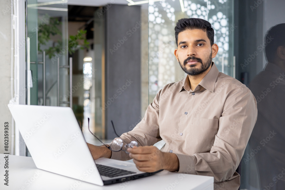 Fototapeta premium Serious islamic businessman taking off glasses during working day in personal workplace with computer. Concentrated startup runner making hard decisions about business growing strategy.