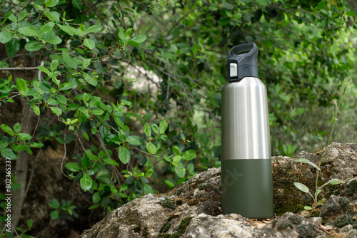 Reusable stainless steel water bottle over a rock surrounded by greenery, ecofriendly lifestyle, space for copy