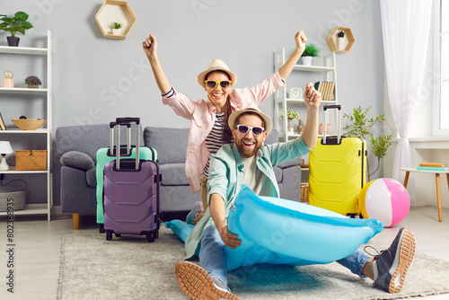 Playful funny couple having fun on air mattress at home, imagining themselves on summer vacation. Man and woman in summer hats and sunglasses are having fun at home among colorful suitcases. photo