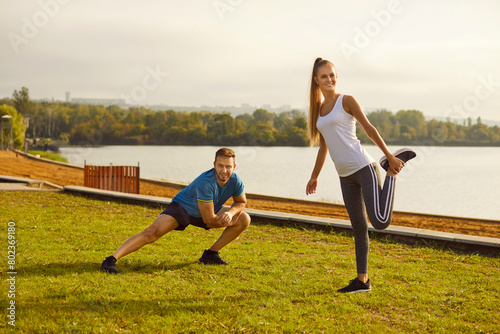 Young fit and active couple doing sport stretching exercising in nature. Happy smiling man and woman in sportswear having workout in the park. Outdoors training and fitness concept.