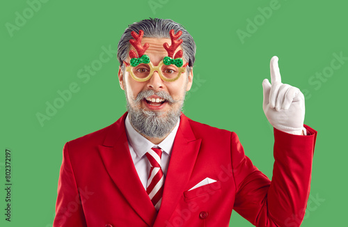 Grey haired bearded man in funny Xmas glasses with antlers, red suit having a good idea pointing index finger up on green background and looking at camera. Christmas, New Year, winter holiday banner. photo