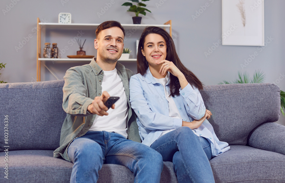 Cozy togetherness. Cheerful couple is relaxing with remote control in their hands, choosing movie or show to watch. Young Caucasian girlfriend and boyfriend sitting together on sofa in front of TV.