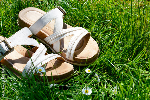 Two white sandals are on the green grass in the middle of the summer day. The green field with two white fashion sandals