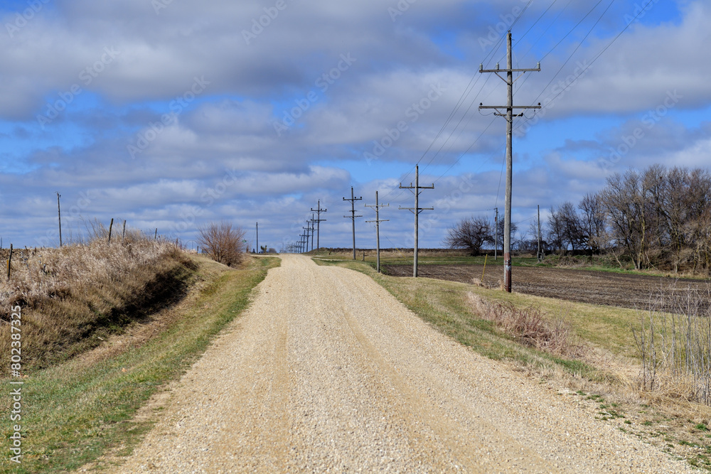 A unpaved country road stretches to the horizon on a sun-drenched late winter day in rural Illinois. The empty roadway sees very little traffic being used predominantly by farm residents in the area. A unpaved country road stretches to the horizon on a sun-drenched late winter day in rural Illinois. The empty roadway sees very little traffic being used predominantly by farm residents in the area.