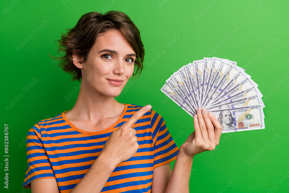 Photo portrait of lovely young lady finger point money fan credit dressed stylish striped garment isolated on green color background Photo portrait of lovely young lady finger point money fan credit dressed stylish striped garment isolated on green color background