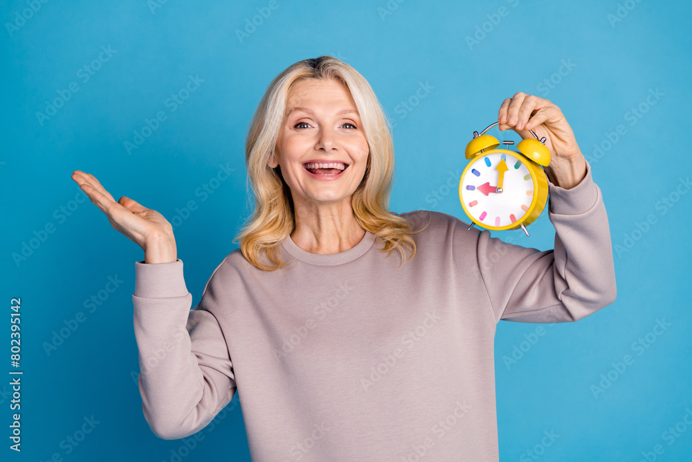 Photo portrait of lovely senior lady hold alarm clock timer dressed stylish gray garment isolated on blue color background Photo portrait of lovely senior lady hold alarm clock timer dressed stylish gray garment isolated on blue color background