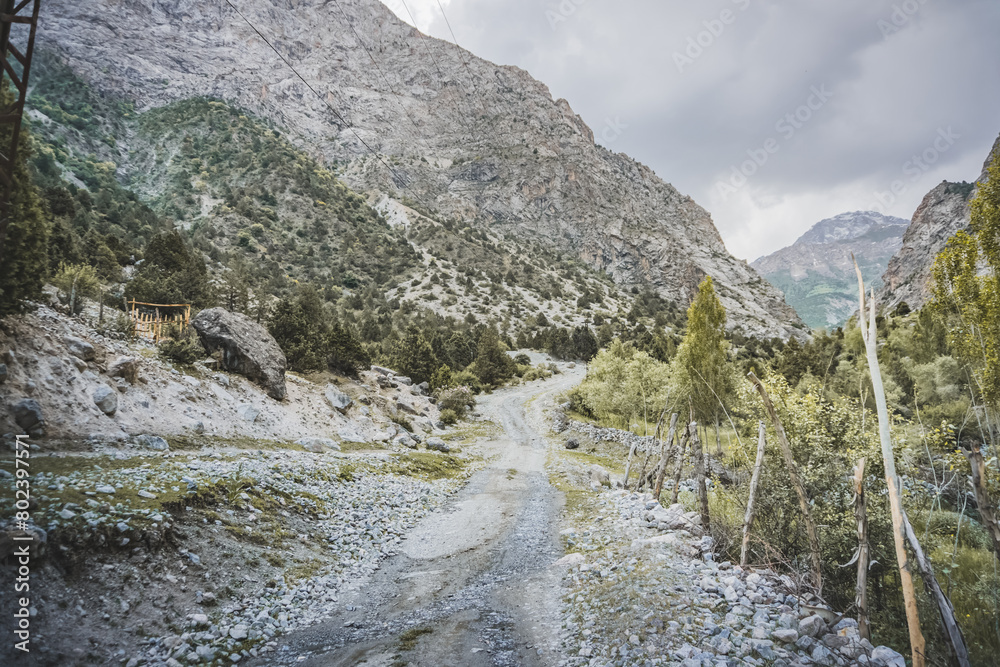 Dusty mountain road in a valley among rocks and vegetation in the Fann Mountains in Tajikistan with mountain ranges, road high in the mountains Dusty mountain road in a valley among rocks and vegetation in the Fann Mountains in Tajikistan with mountain ranges, road high in the mountains