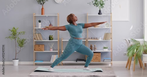 Yogi African American chubby woman doing yoga at home, standing in Warrior one exercise, nice lady losing weight, pilates, working out, wearing sportswear, performing Virabhadrasana I pose photo