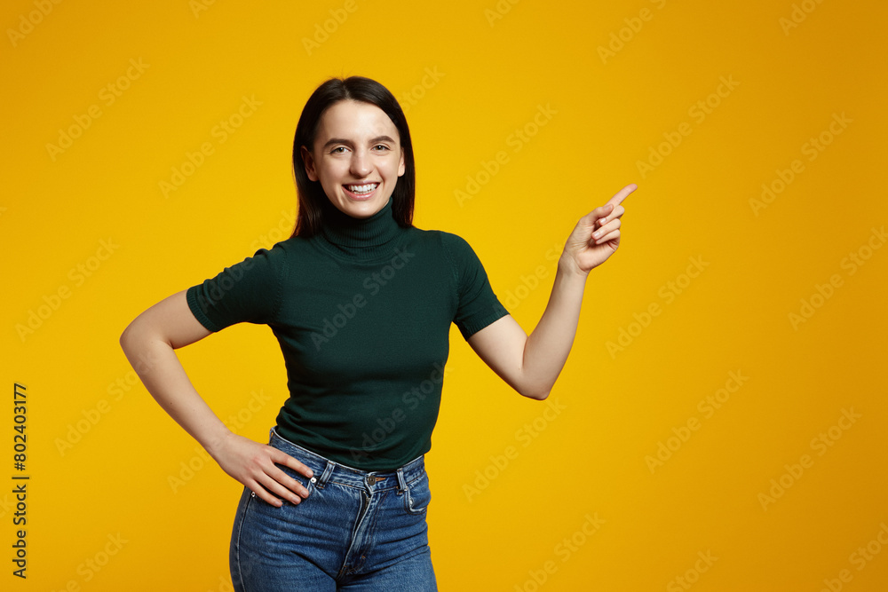 Young confident woman showing logo on empty space. Smiling girl inviting or advertising promo deal, pointing fingers left and looking at camera, yellow background Young confident woman showing logo on empty space. Smiling girl inviting or advertising promo deal, pointing fingers left and looking at camera, yellow background