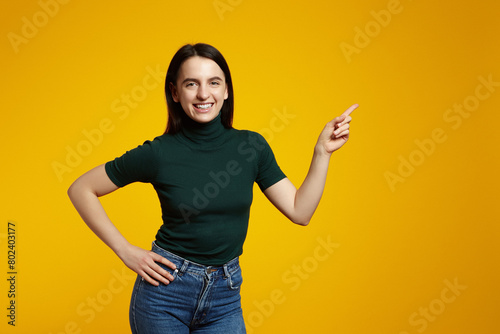 Young confident woman showing logo on empty space. Smiling girl inviting or advertising promo deal, pointing fingers left and looking at camera, yellow background