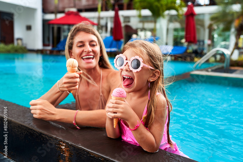 Cheerful mother and daughter enjoy ice cream by poolside. Little girl in pink swimsuit, goggles eats dessert, laughs. Mom joins fun on sunny day. Family pool time, sweet treat moment captured.