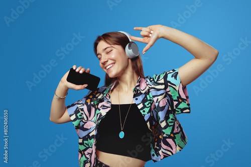 Cheerful young girl in headphones smiling and showing rock gesture while holding smartphone with closed eyes, isolated against blue background. Sign, symbols concept