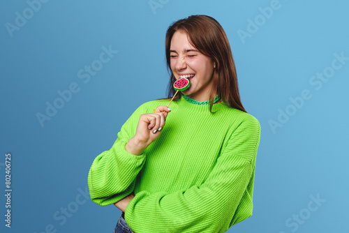 Young pretty lady in bright green sweater keeping eyes closed and smiling while biting lollipop, standing isolated over blue background.