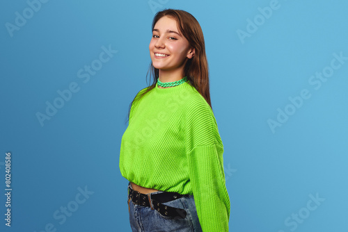 Cute young girl in trendy outfit standing with hands behind back and looking at camera against blue background.