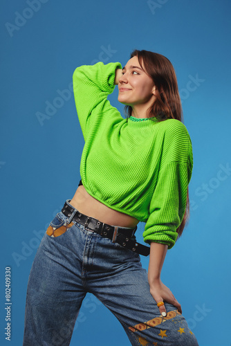 Photo of glad young girl in trendy outfit with long brown hair, holding hand behind head and looking away with smile while standing against blue background.