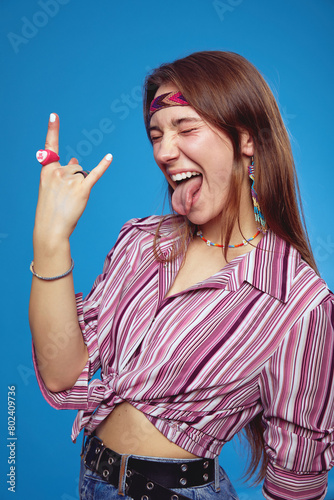 Vertical photo of young excited hippie woman makes rock n roll gesture with hand up, showing tongue with closed eyes, isolated over blue background
