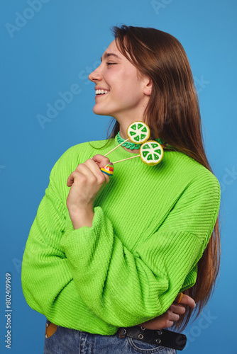 Vertical side view portrait of cheerful adolescent girl smiling and holding two lemon candy on stick, wearing bright green sweater and looking away, isolated over blue background.