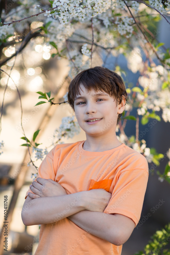 Happy preteen boy wearing an orange T-shirt Looks at looks at the camera in a blossoming garden. Happy preteen boy wearing an orange T-shirt Looks at looks at the camera in a blossoming garden.