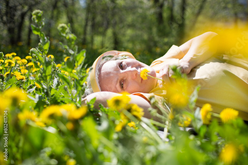  lifestyle portrait senior woman  with gray hair in headphones relaxeslying on the grass with dandelions in spring. Audio healing. meditation. banner. copy space. 