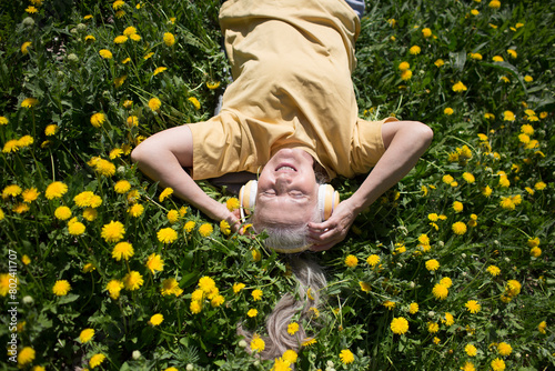  lifestyle portrait senior woman  with gray hair in headphones relaxeslying on the grass with dandelions in spring. Audio healing. meditation. banner. copy space. 