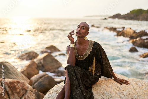 Non-binary black person in luxury dress sits on rocks in ocean. Trans ethnic fashion model wearing jewelry in posh gown, poses in tropical seaside location. Divine feminine human. Diversity concept. photo