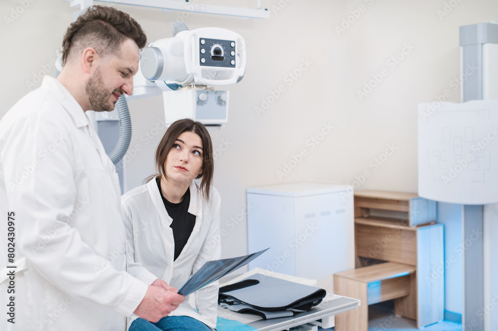 A male doctor shows xray photo to female patient to take an x-ray on a special stand in an office with an x-ray machine, medical equipment. A male doctor shows xray photo to female patient to take an x-ray on a special stand in an office with an x-ray machine, medical equipment.